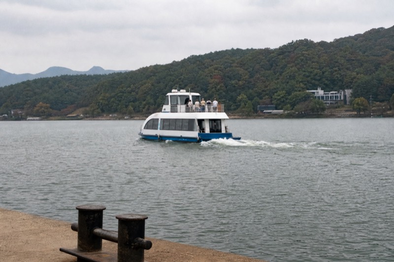 A small passenger ferry crossing a wide calm river toward a tree-covered island, the wooded bank of Gapyeong behind it